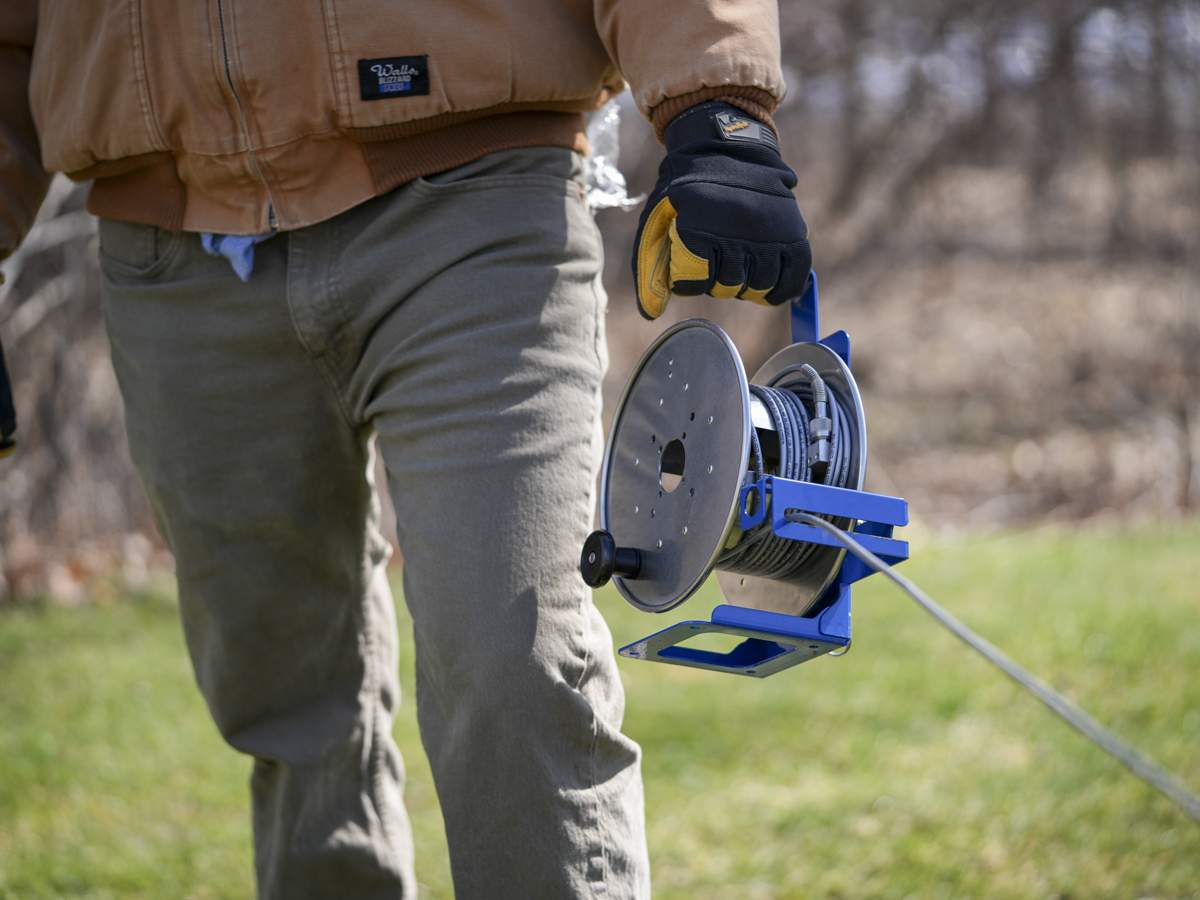 Technician carrying the QTHR Hose Reel Kit with pressurized hose for remote transmitter calibration.